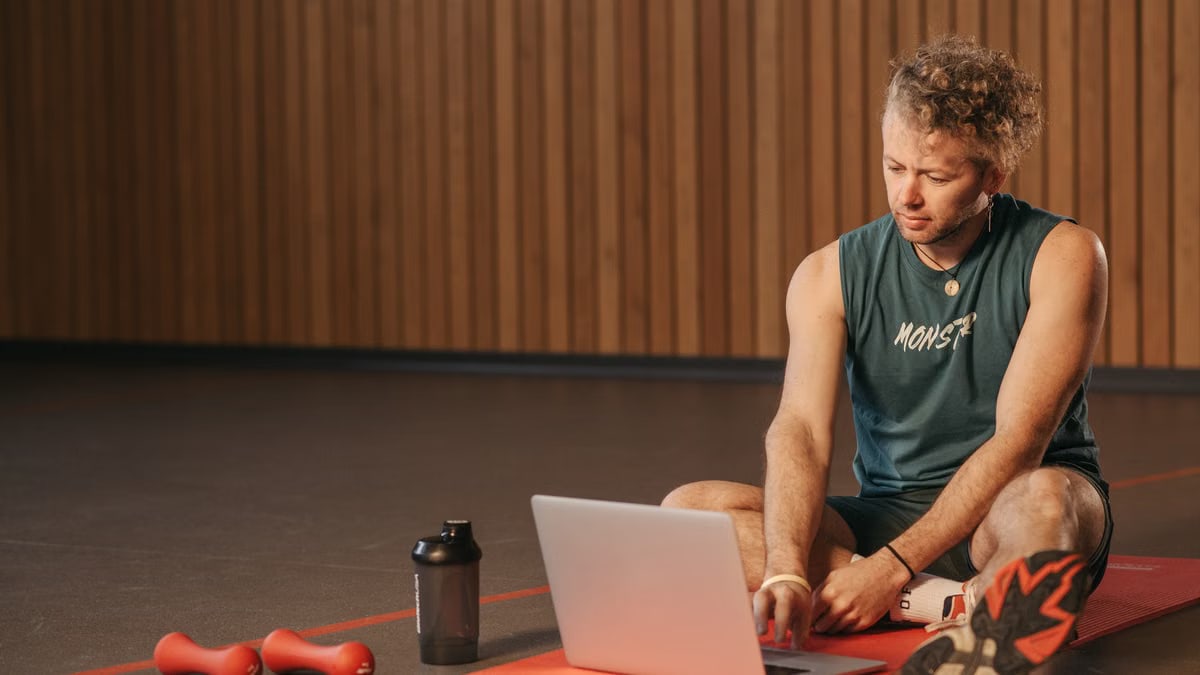 Man sitting on exercise mat with laptop, surrounded by dumbbells, creates a unique fitness workspace.