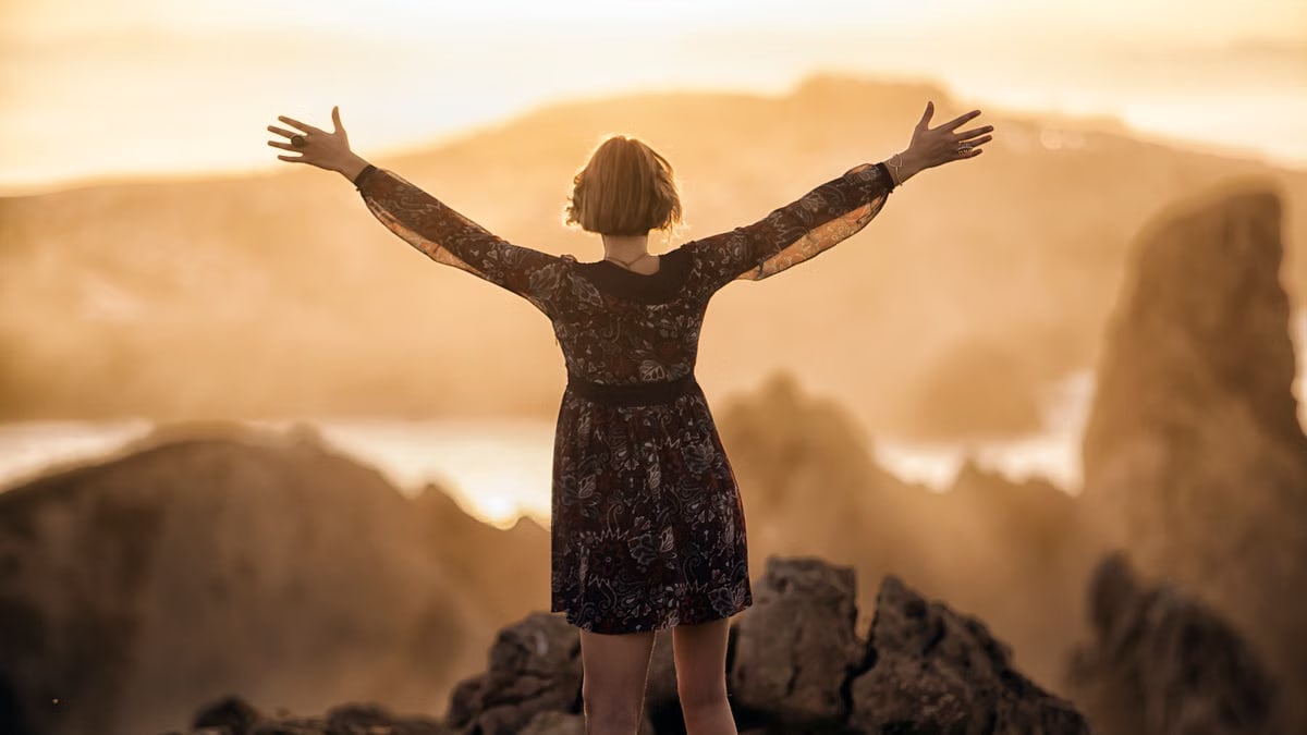 A woman stands with open arms on a mountain at sunset, embracing nature's beauty.